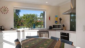 A modern kitchen with white cabinets, a dining table with a patterned table runner, and a window overlooking greenery. - The Peninsula Riverside Abode with Garden Views (Auckland West)