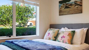 A bedroom with a large window, a bed with floral pillows, and a painting of boats on the wall. - The Peninsula Riverside Abode with Garden Views (Auckland West)