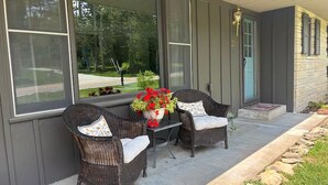 A porch with wicker chairs and a table, a potted plant, and a view of a road and trees. - Stadium Stroll Main Level | Walk to Football | Sleeps 8 · 2 BR · 1 Bath (Bloomington)