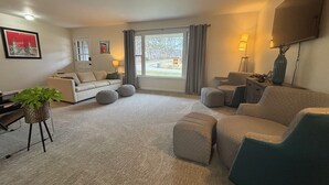 A modern living room with a sofa, ottomans, a TV, and a large window with curtains. - Stadium Stroll Main Level | Walk to Football | Sleeps 8 · 2 BR · 1 Bath (Bloomington)