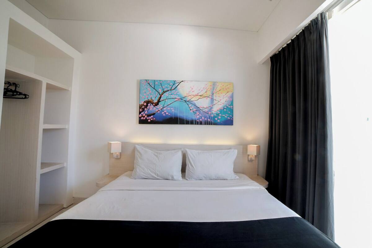 A hotel room with a bed, two bedside lamps, a wall-mounted painting, and a wardrobe.