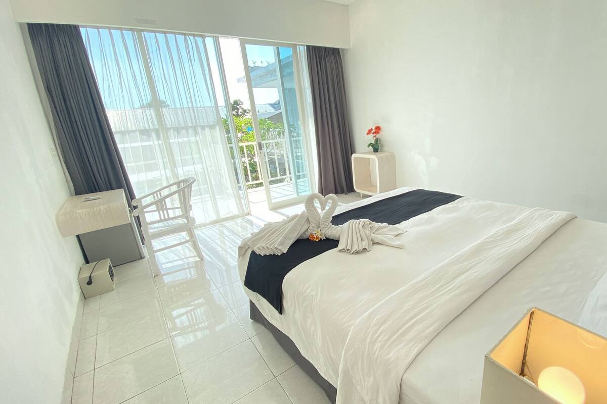 A hotel room with a large bed, a bedside table, a chair, a desk, and a view of the outside.