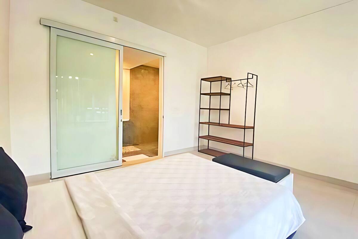 A modern bedroom with a sliding glass door leading to a bathroom, a built-in bench, and a metal shelving unit.