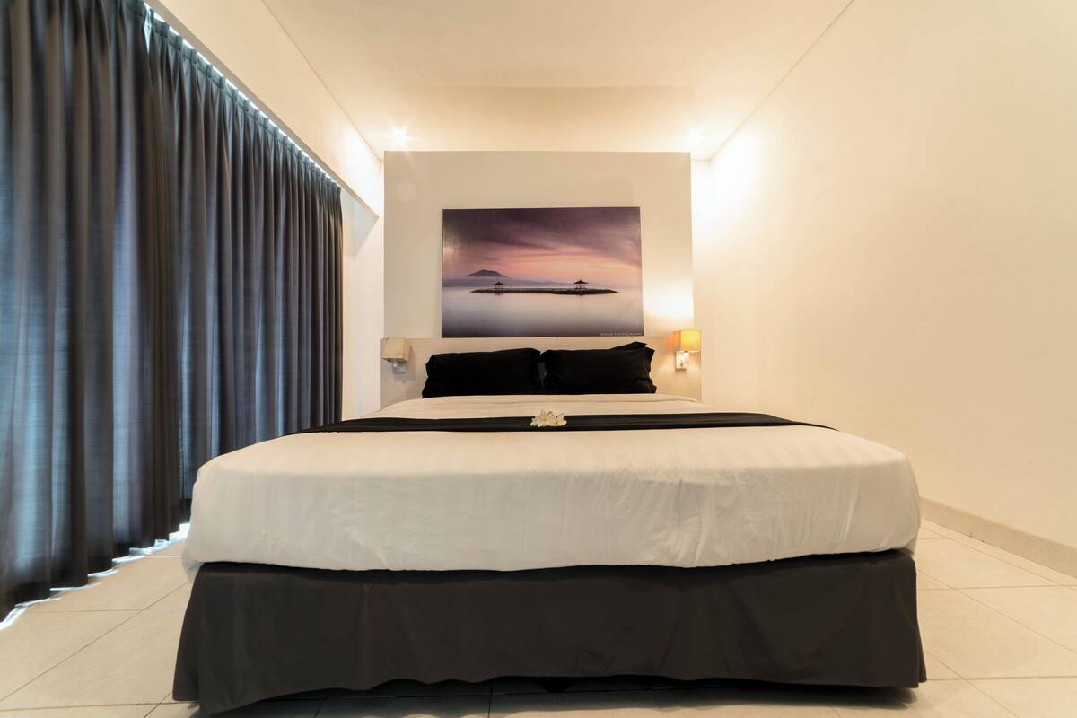 A hotel room with a large bed, a painting of a sunset over water, and a view of the sea through the window.