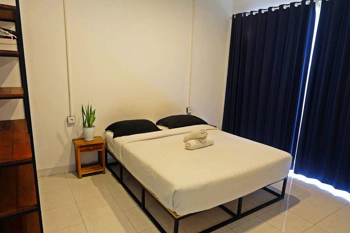 A single bed with a white mattress and black headboard, a small wooden nightstand with a potted plant, and a dark curtain covering a window.