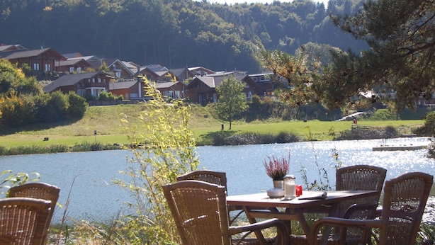 A lakeside view with a dining table and chairs, surrounded by houses and a forested hill. - Vacation home Holunder - 7 min. walk to the bathing lake Waldsee Rieden (Rieden)