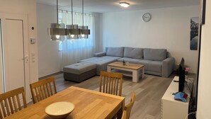 A modern living room with a grey sofa, a wooden dining table, and a kitchen area with a computer and a plant. - Vacation home Holunder - 7 min. walk to the bathing lake Waldsee Rieden (Rieden)