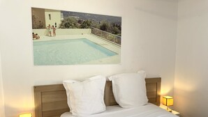 Un dormitorio con cama, almohadas blancas, cabecera de madera y un cuadro enmarcado con una piscina en la pared.