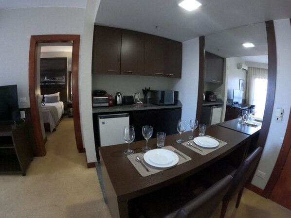 Habitación de hotel pequeña con kitchenette, un comedor con una mesa dispuesta para dos personas, y un dormitorio separado visible a través de una puerta abierta.