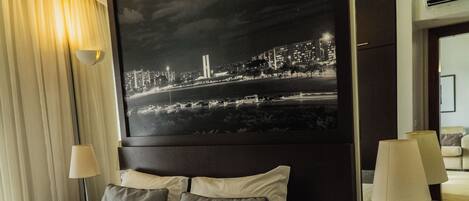 A hotel room with a neatly made bed, two bedside lamps, a nightstand with a phone, and a large cityscape painting on the wall.