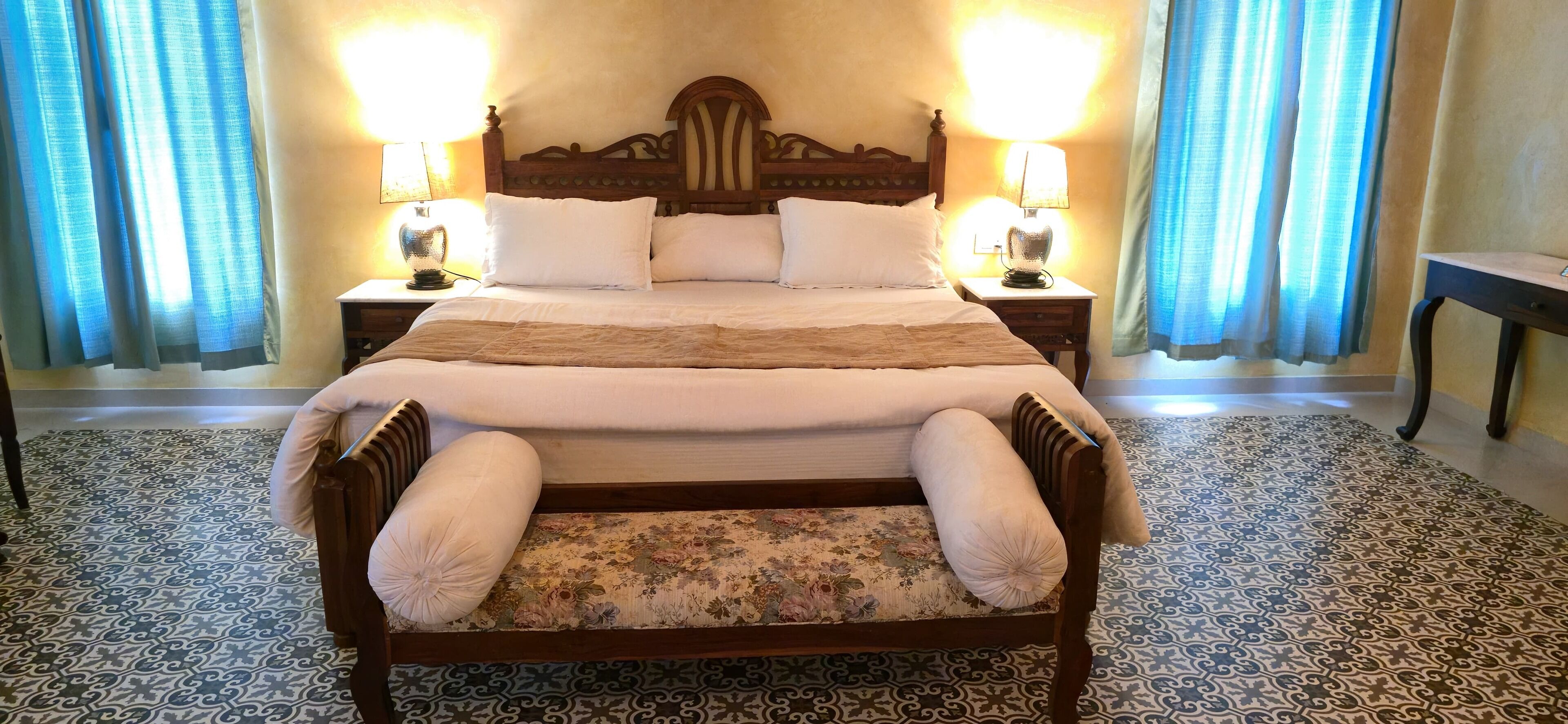 A large bed with a wooden headboard, two bedside tables with lamps, and a floral-patterned bench.