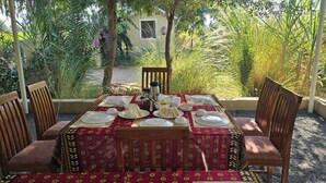 Un espace extérieur aménagé pour les repas, avec une table dressée pour quatre personnes, entourée de chaises, et une vue sur un jardin où l’on peut admirer différentes plantes, ainsi que sur un bâtiment en arrière-plan.