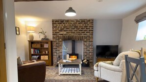 A cozy living room with a brick fireplace, a television, a sofa, and a chair. - Traditional Country Cottage (Pennant)