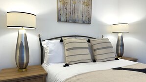 A neatly made bed with two bedside lamps, a headboard, and a painting above it. - Traditional Country Cottage (Pennant)