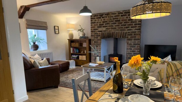 A cozy living room with a fireplace, a dining table set for two, and a comfortable sofa. - Traditional Country Cottage (Pennant)