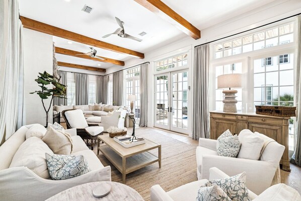 A spacious living room with a high ceiling, wooden beams, a ceiling fan, and large windows.