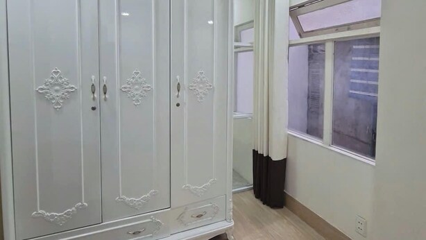 Ein geräumiges Zimmer mit großem weißen Kleiderschrank, einem fenster mit Vorhängen und einer Decke mit Einbuchtungslampen.