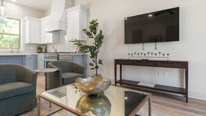 A modern living room with a glass coffee table, a sofa, a TV, and a kitchen in the background.
