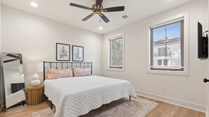 A bedroom with a bed, a ceiling fan, a window with blinds, a TV mounted on the wall, and a bedside table with a lamp.