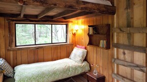 A rustic cabin bedroom with a bunk bed, a window with a view of the forest, a bedside lamp, and a small bookshelf. - Outlier Inn Tiny House 4 on Farm Upstate Catskills (Woodridge)