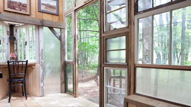 A rustic wooden room with large windows, a chair, and a small table. - Outlier Inn Tiny House 4 on Farm Upstate Catskills (Woodridge)