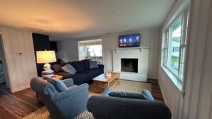A living room with a fireplace, a television mounted above it, and a dining area with a table and chairs. -  Charming Cottage w/ WiFi, AC in York, Walking Distance to Long Sands Beach (York)