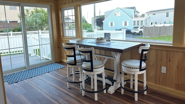 A room with a wooden paneled wall, a large window, a table with four chairs, and a view of a residential area. -  Charming Cottage w/ WiFi, AC in York, Walking Distance to Long Sands Beach (York)