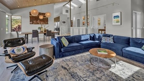 A spacious living room with a blue sectional sofa, a leather armchair, and a coffee table. There is a dining area with a wooden table and chairs, and a kitchen with modern appliances. The room has large windows allowing natural light and a view of the outdoors.