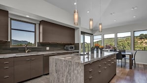 A modern kitchen with a large island, dark cabinetry, and large windows offering a view of the outdoors.