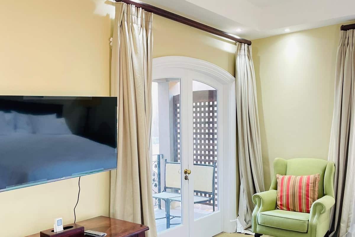 A hotel room with a flat-screen TV mounted on the wall, a wooden cabinet with electronic devices, a green armchair with a striped pillow, and a door leading to an outdoor area with a table and chairs.