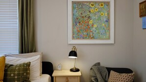 A room with a bed, a nightstand with a lamp, a framed floral painting, and a window with curtains.