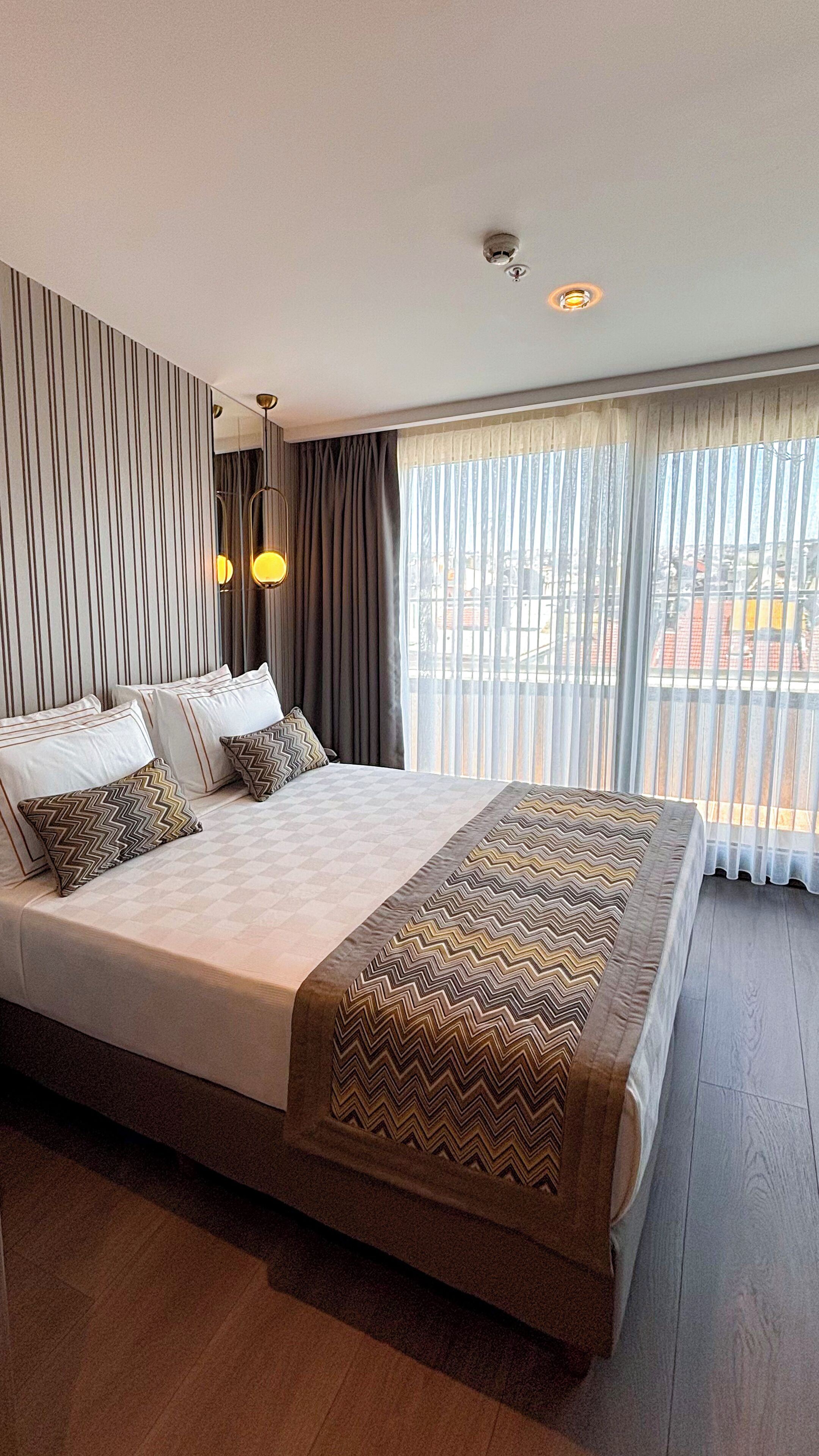 A hotel room with a large bed, a mirror, a window with curtains, and a ceiling light fixture.