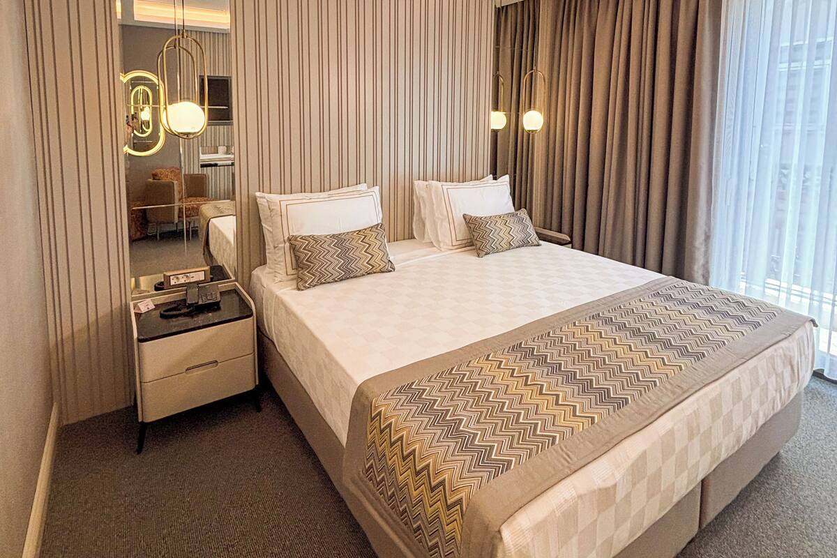 Habitación de hotel con una cama grande, dos mesitas de noche, un espejo y vistas al exterior.