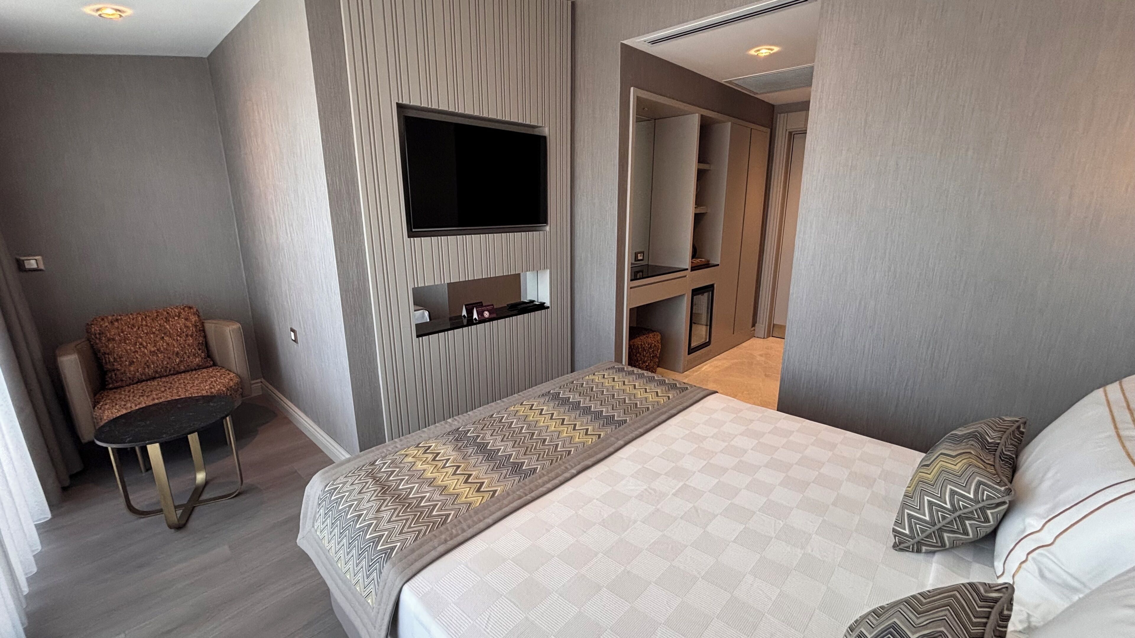 A modern hotel room with a bed, a small seating area, a TV mounted on the wall, and a closet with built-in shelves.