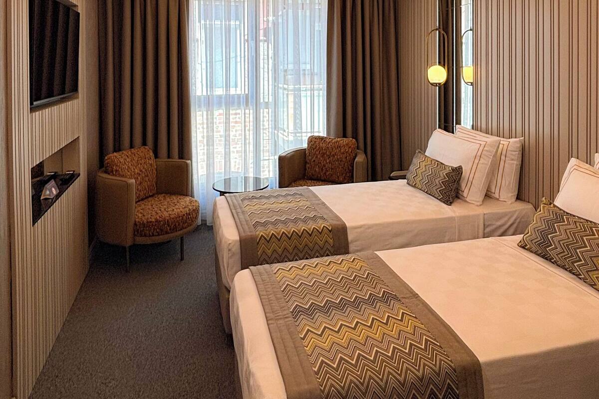 Habitación de hotel con dos camas, televisión, zona de estar y un amplio ventanal con cortinas.