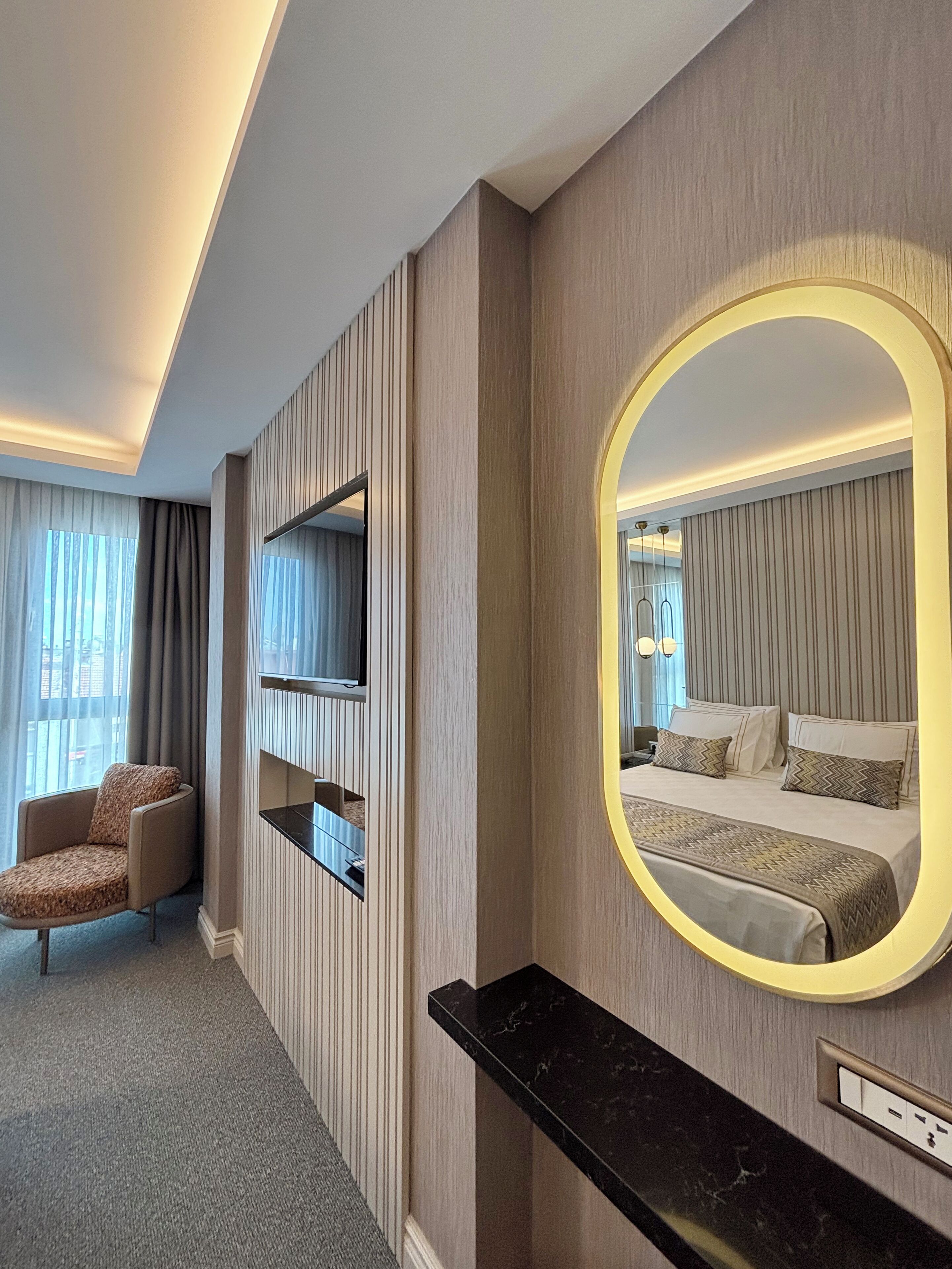 A hotel room with a bed, a TV mounted on the wall, a chair, and a large mirror with a gold frame.