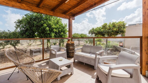 Une terrasse couverte, aménagée avec des meubles en osier, une table en verre et offrant une vue sur la verdure environnante.