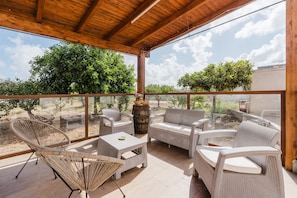 Un patio coperto con mobili in vimini, un tavolo in vetro e vista sul verde circostante.