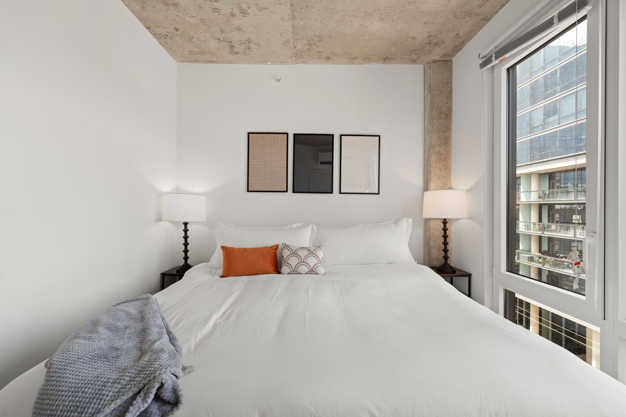 A modern hotel room with a large bed, two bedside tables with lamps, a large window with a view of a building, and three framed artworks on the wall.