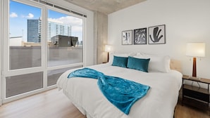 Une chambre d’hôtel moderne dotée d’un grand lit, offrant une vue sur le paysage urbain grâce à de grandes fenêtres du sol au plafond, et ornée de trois œuvres d’art encadrées au mur.