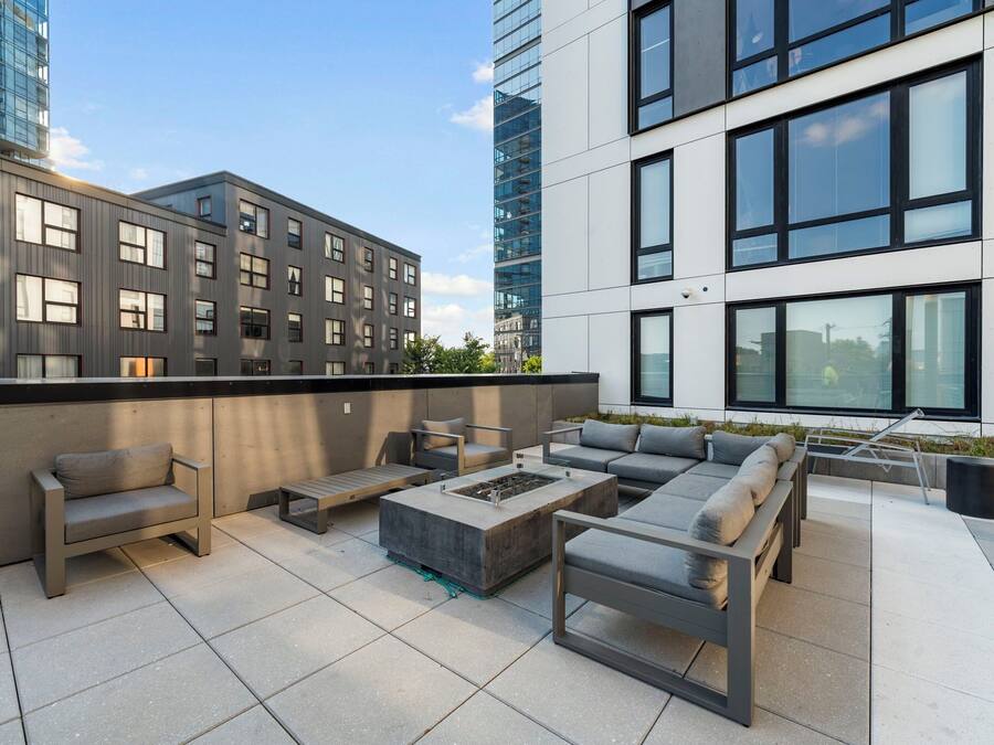 An outdoor terrace with modern furniture, including sofas and a fire pit, surrounded by buildings with large windows and a clear sky.