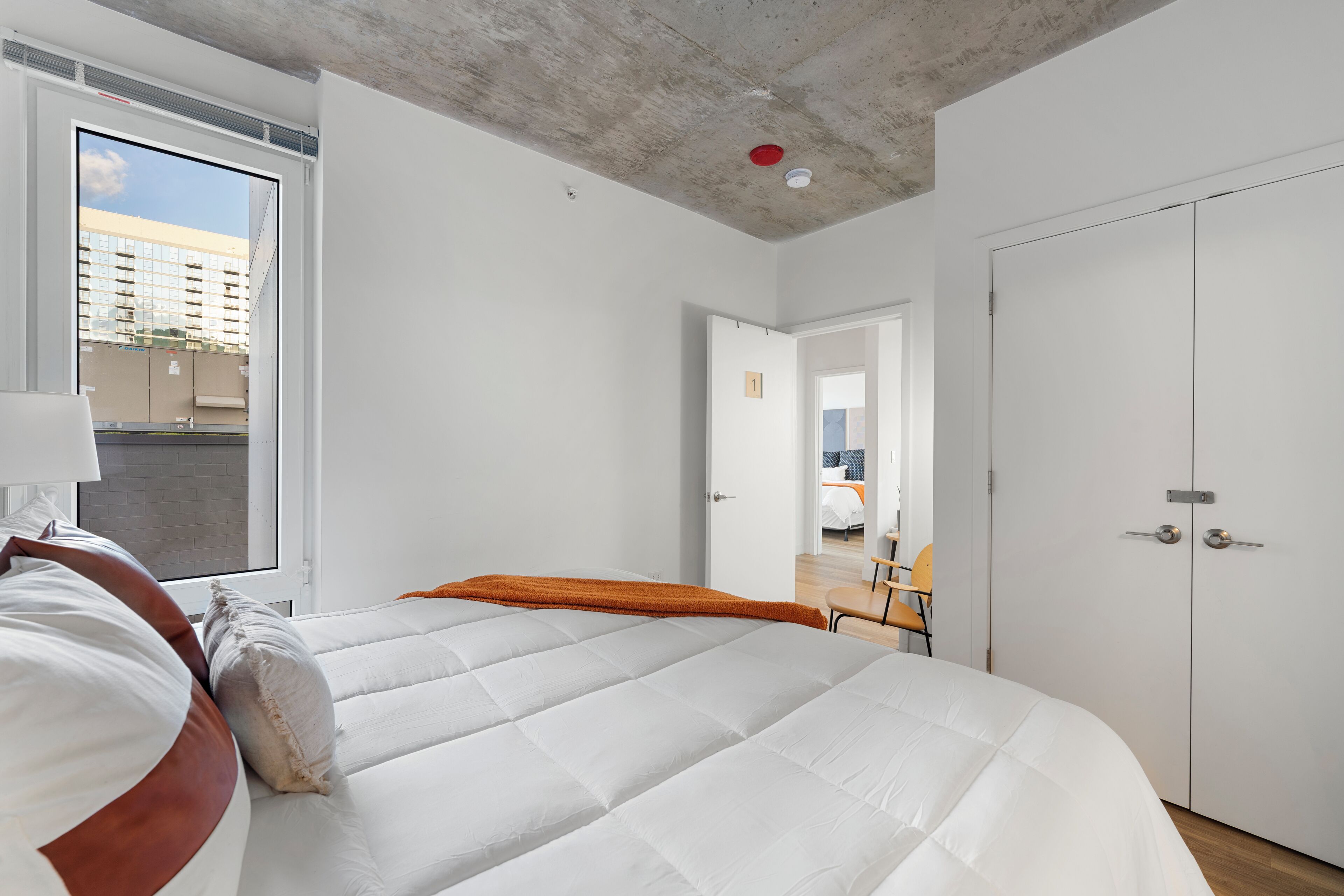 Een moderne slaapkamer met een groot bed, een raam met uitzicht op een gebouw en een deur naar een andere kamer.