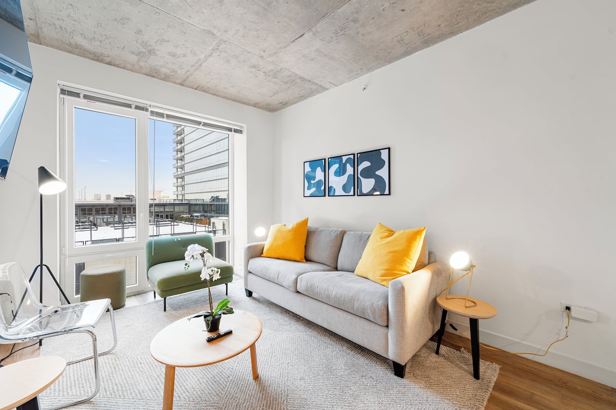 A modern living room with a grey sofa, yellow cushions, a small round table, and a green armchair. There are three abstract paintings on the wall, a large window with a view of buildings, and a ceiling with exposed concrete.