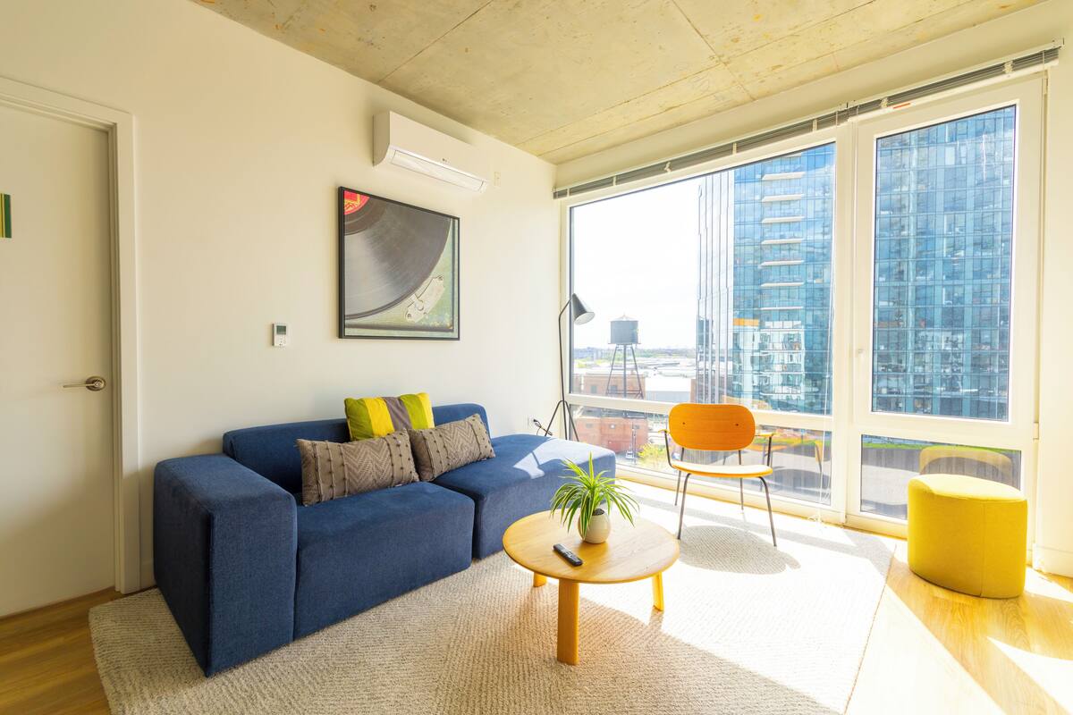 A modern living room with a blue sofa, a round coffee table with a plant, a yellow ottoman, and a large window with a city view.