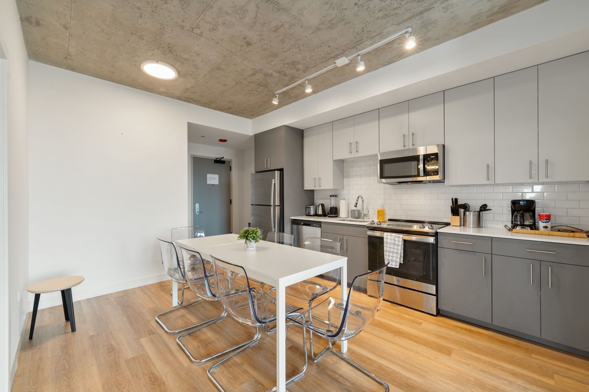 A modern kitchen with grey cabinets, stainless steel appliances, and a white dining table with clear chairs.