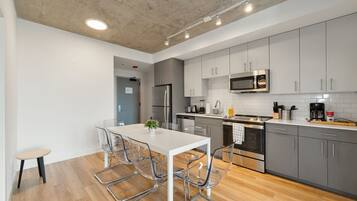 A modern kitchen with grey cabinets, stainless steel appliances, and a white dining table with clear chairs.