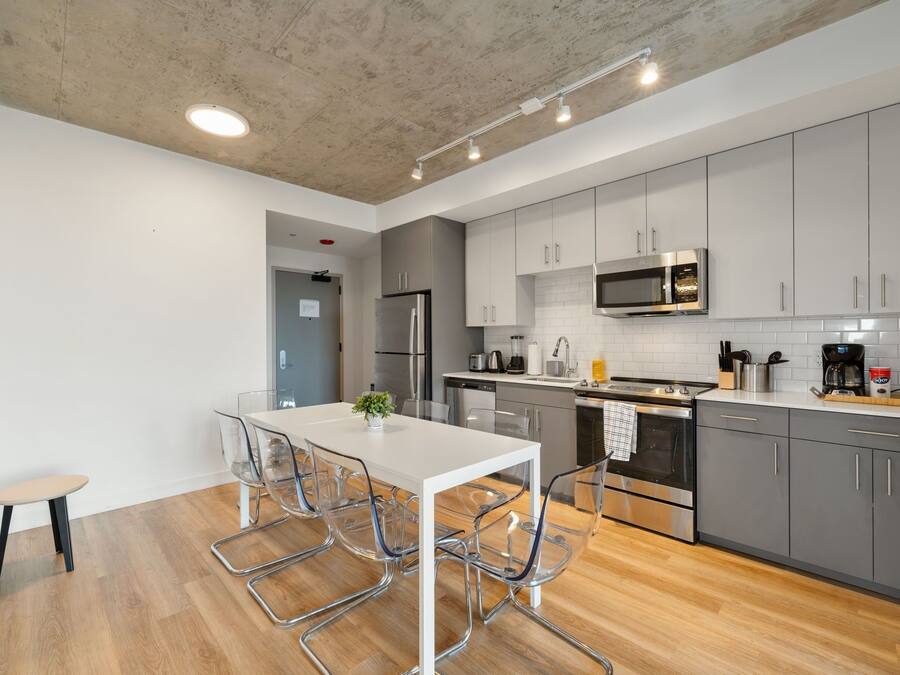A modern kitchen with grey cabinets, stainless steel appliances, and a white dining table with clear chairs.