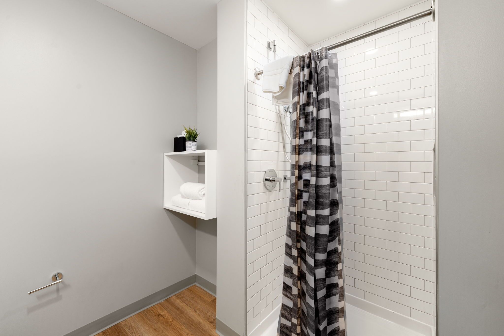 Un baño moderno con azulejos blancos tipo metro, ducha con cortina a rayas negras y blancas, un estante con una planta y un perchero.