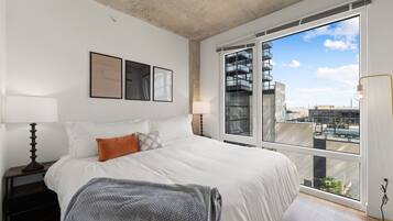 Un dormitorio con un ventanal grande, una cama con ropa blanca, dos lámparas fijadas a la pared y obras de arte enmarcadas.