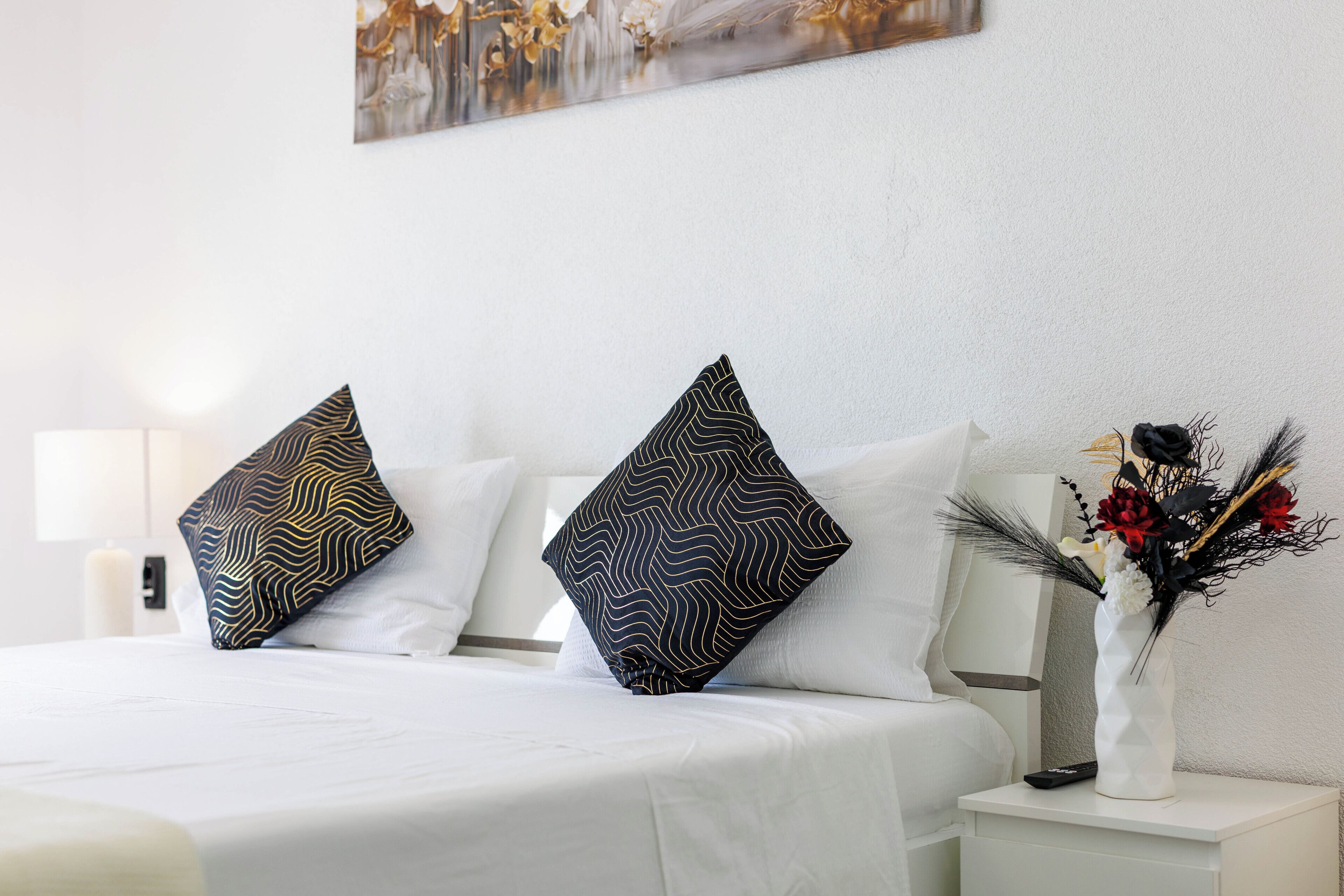 A neatly made bed with patterned pillows, a white bedside table with a vase of flowers, and a painting on the wall.